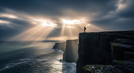 Solitary figure stands silhouetted on dramatic coastal precipice beneath radiant sunbeams piercing stormy clouds