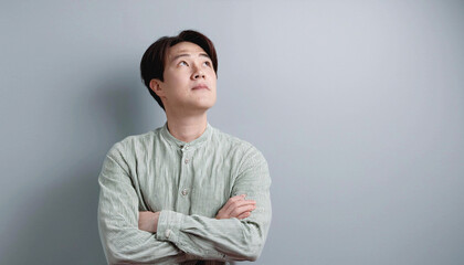 グレーの壁を背景に、腕を組む日本人男性。左側に男性が寄っているのコピースペースが広い。男性は上を斜め上を見上げている。モスグリーンのラフなシャツを着ている。