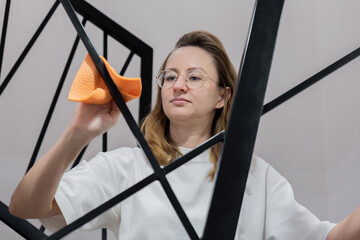 young woman cleaning glass mirror with orange cloth, wearing white shirt and glasses, focused expression, wiping black geometric frame, close-up interior scene, performing routine home maintenance