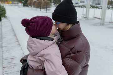 mother leaning in whispering to daughter on snow-lined path, daughter reacting with giggle and...