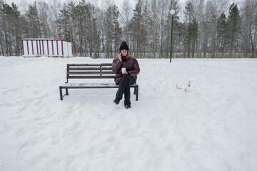 Fototapeta premium solitary woman sitting on snowy bench gazing into distance, wide winter landscape with sparse trees and modern building backdrop, mood of contemplation and quiet pause, footprints in snow and muted