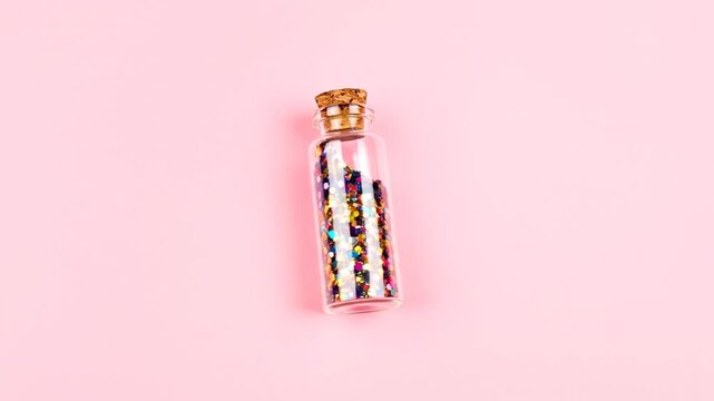 A clear glass bottle with a cork stopper is filled with colorful glitter on a pink background
