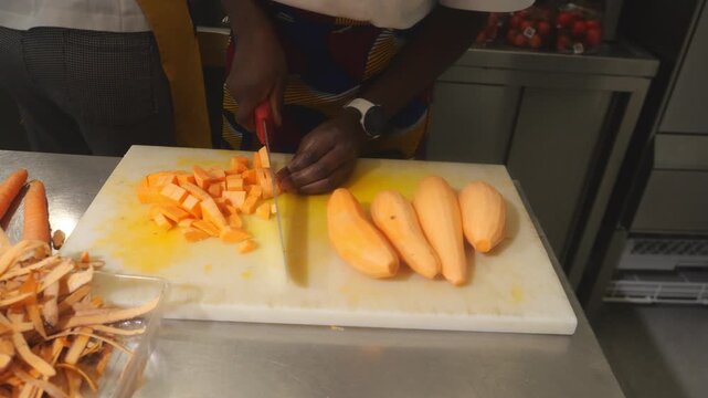 Chef in a vibrant patterned apron and white digital watch uses a stainless steel knife to peel and dice orange-fleshed yams Ipomoea batatas with a Y-peeler and then cut into precise uniform concass&eacute;