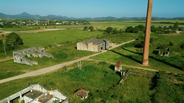 Low flying drone moves forward over an abandoned house toward at Santa Luiza sugar and ethanol mill in Saquarema, Brazil. Industrial ruins, green fields and twin chimneys dominate the rural landscape