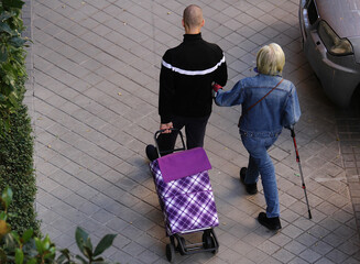 Hijo con madre agarrada del brazo con bast&oacute;n y carro en perspectiva a&eacute;rea