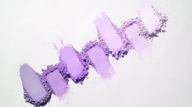 Top view shot of Seven violet powder smears arranged in a row on a pale gray canvas. Dynamic orbiting light projects moving shadows