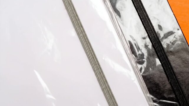 Two notebooks, one white, one black, sit on an orange surface. Both are sealed in clear plastic