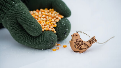 A close-up shot of hands in knitted mittens placing a handful of grains on the snow, with a small wooden amulet in the shape of a bird next to them.