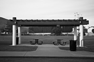 Open park pavilion with picnic tables and shaded seating, set on a grassy field with mountains and trees in the background.