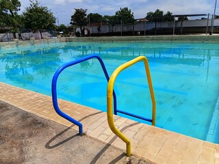 Yellow and blue metal handrails at the edge of a public swimming pool. Close-up of pool entry with clear water and tan tiles, ideal for summer recreation and safety concepts.