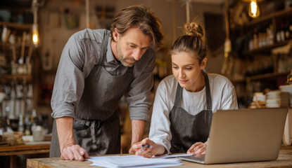 A man and woman in casual aprons collaborating in a cozy kitchen, reviewing recipes or food preparation plans together at a rustic wooden table