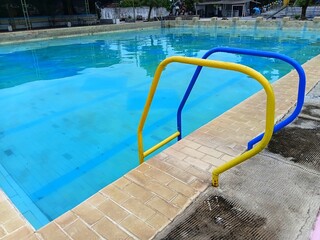 Yellow and blue metal handrails at the edge of a public swimming pool. Close-up of pool entry with clear water and tan tiles, ideal for summer recreation and safety concepts.