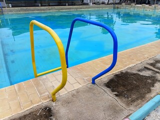 Yellow and blue metal handrails at the edge of a public swimming pool. Close-up of pool entry with clear water and tan tiles, ideal for summer recreation and safety concepts.
