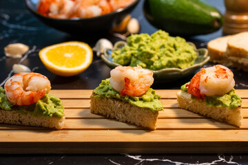 three shrimp and avocado toast appetizers resting on a wooden slat board, with a bowl of guacamole...