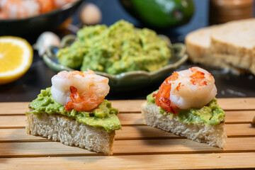 two shrimp and avocado toast appetizers resting on a wooden slat board, with a bowl of guacamole and fresh lemon blurred in the background.