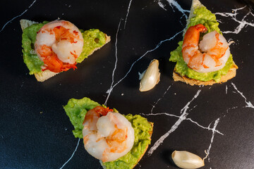 flat lay showing three triangular toast appetizers topped with creamy avocado spread and cooked shrimp, arranged on a black marble-patterned surface with scattered garlic cloves.