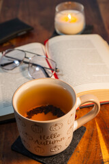 a cozy reading setup featuring an open book with glasses, a steaming mug of tea in the foreground, and a lit candle on a wooden table.