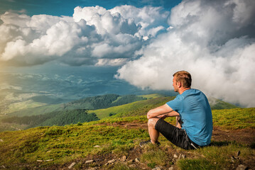 Man is sitting on the grass in the mountains and is looking on the stormy clouds