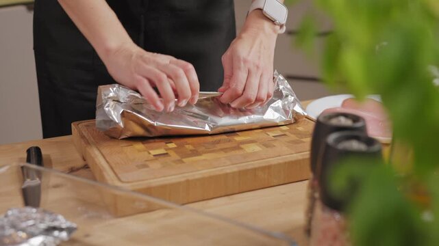 Close-up of woman hands wrapping seasoned raw chicken breast in aluminum foil on wooden cutting board in kitchen. Concept of food preparation and cooking process