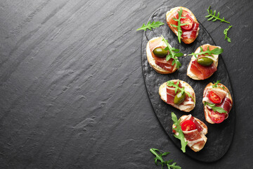 Delicious bruschettas with cream cheese, prosciutto, olives, tomatoes and arugula on dark textured table, top view. Space for text