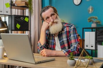 Thoughtful concerned Caucasian senior man looking away thinking solving problem with laptop at desk in home office. Serious grandpa freelancer search for inspiration make decision feel lack of ideas.