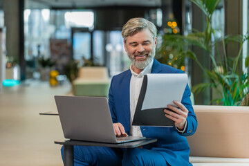 Happy businessman using laptop and working with documents, analyzing information, reading papers prepare financial report. Male executive in suit closing netbook after finishing work in office lobby.