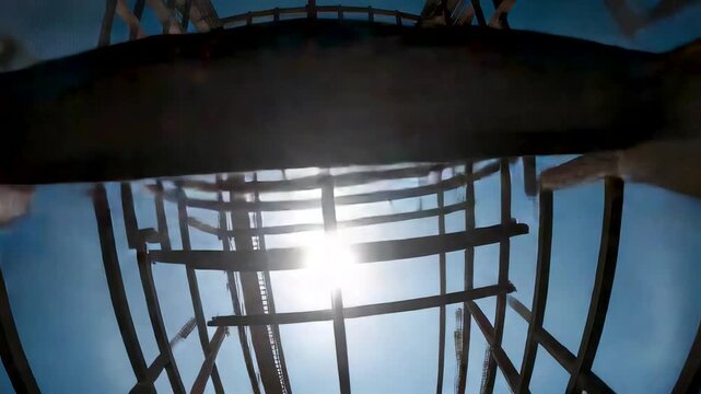 Upward perspective of steel tower framework with sunlight flare and curved metal elements forming abstract industrial architecture against blue sky