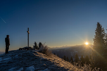 mountain summit cross and hiker in winter towards sunrise above the summits of Bregenzerwald in Vorarlberg Austria on a clear frosty morning 