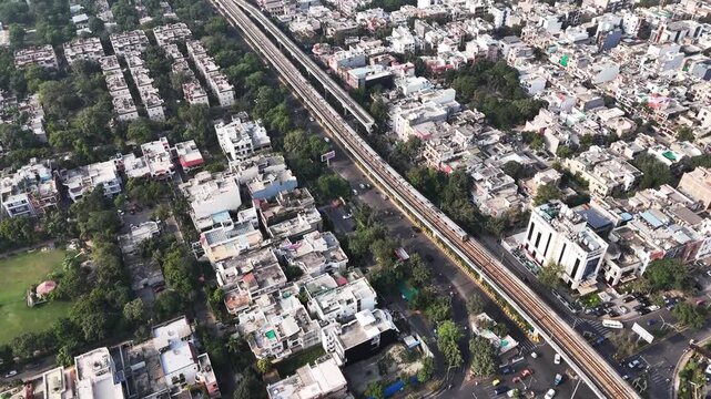 High-angle aerial of Noida, elevated metro train moving on a track spanning across traffic-filled roads and residential areas, everyday city movement and modern transport shaping the NCR landscape.
