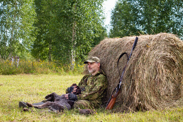 A hunter hugs his dog while relaxing in the shade of a hay bale