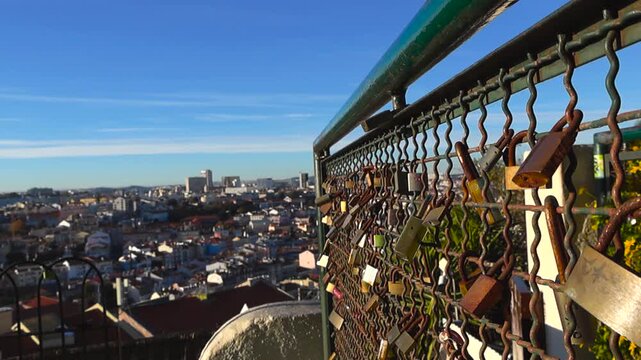 Smooth glide along a railing covered with love padlocks at Miradouro do Jardim do Torel with a wide sunny view of Lisbon rooftops and the Tagus River in the background.