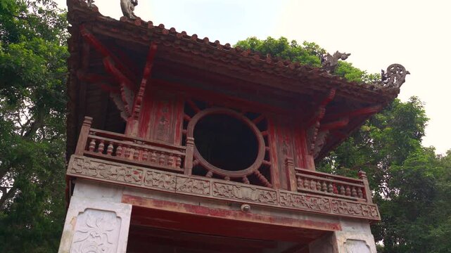 Low angle of Khue Van Cac Pavilion at the Temple of Literature in Hanoi. Iconic red wooden architecture with circular window surrounded by green trees in a historic Vietnamese site.