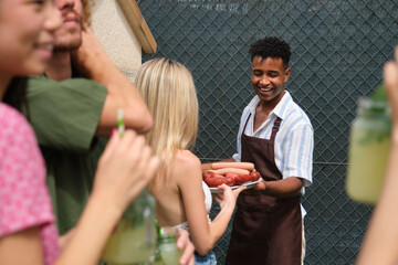 Waiter serving grilled sausages to friends drinking cocktails at barbecue party