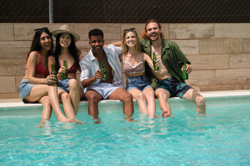 Happy young people sitting at the poolside are holding bottles and smiling