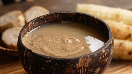A refreshing traditional beverage possibly kava or a root drink being carefully poured into a rustic coconut shell bowl with blurred taro roots and sugarcane stalks visible in the background evoking .
