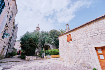 Historic stone courtyard and narrow street with trees and a church tower, Mediterranean architecture under a bright spring sky.
