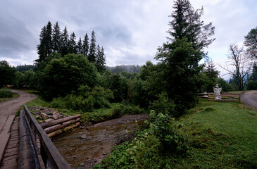 Obraz premium A rustic wooden bridge crosses a small stream in a lush forest landscape under a cloudy sky.
