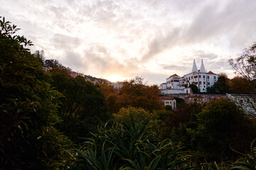 Sintra National Palace and hillside houses glowing at sunset above autumn trees and dense gardens under a dramatic sky in Portugal.