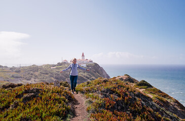 A person walking on a coastal path with a lighthouse in the background, surrounded by ocean and...