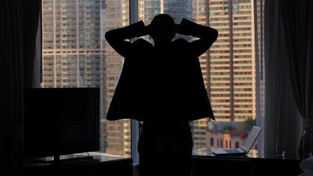 Man in casual suit come to window, put hands on back of head, silhouetted half length shot. Large modern residential building seen blurred outside. Open laptop computer on table