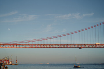 Obraz premium A red suspension bridge spans across the image with a clear blue sky and visible moon.