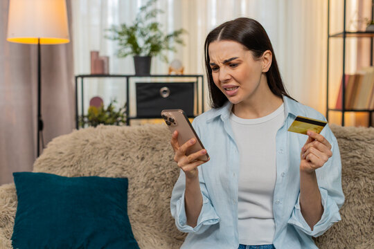 Young woman at home anxiously checking credit card balance on smartphone worried by blocked account or error. Caucasian girl on sofa stressed over financial issues, loss or unsuccessful payment