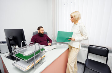 Bright, modern office scene showing a woman with a green folder engaging with a seated man at a desk. They discuss documents in a professional, collaborative environment.