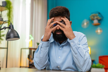 Indian man at home buries face in palms fatigued by burnout stress hopeless after brutal day. Arabian guy at desk shakes quietly feeling deep despair seeking relief from pressure and mental overload.