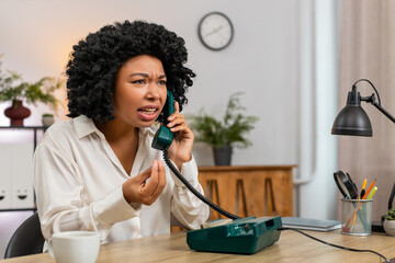 African American businesswoman at home office shouts into retro phone angrily raising voice loud. Freelancer girl at table waves hand expressing urgent frustration demanding quick fix during dispute.
