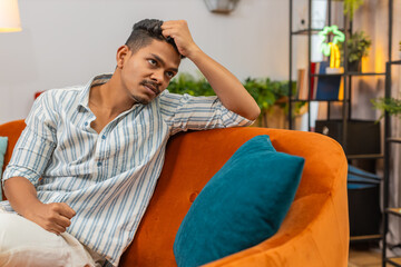 Indian man at home looks down after hard day feels tired low mood with stress and fatigue asks for rest. Arabian guy on sofa sits quiet rubs temples closes eyes and breathes slow to calm and recover.