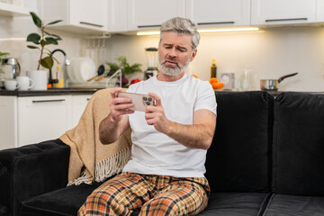 Middle-aged man at home loses play online game on smartphone and sees lottery app bad result sighs. Mature guy on sofa hits retry hopes for luck casino bet failed breath heavy hands tremble gaze down.