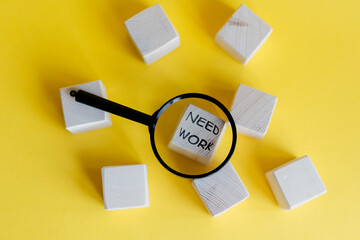 Wooden blocks with the text 'NEED WORK' magnified by a black magnifying glass on a bright yellow background. The scene emphasizes job searching and motivation.