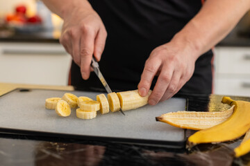 Young man drops sliced banana into blender in home kitchen making smoothie or protein shake for...