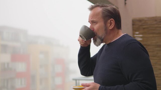A handsome middle-aged man drinks a cup of coffee and looks out a balcony of an apartment